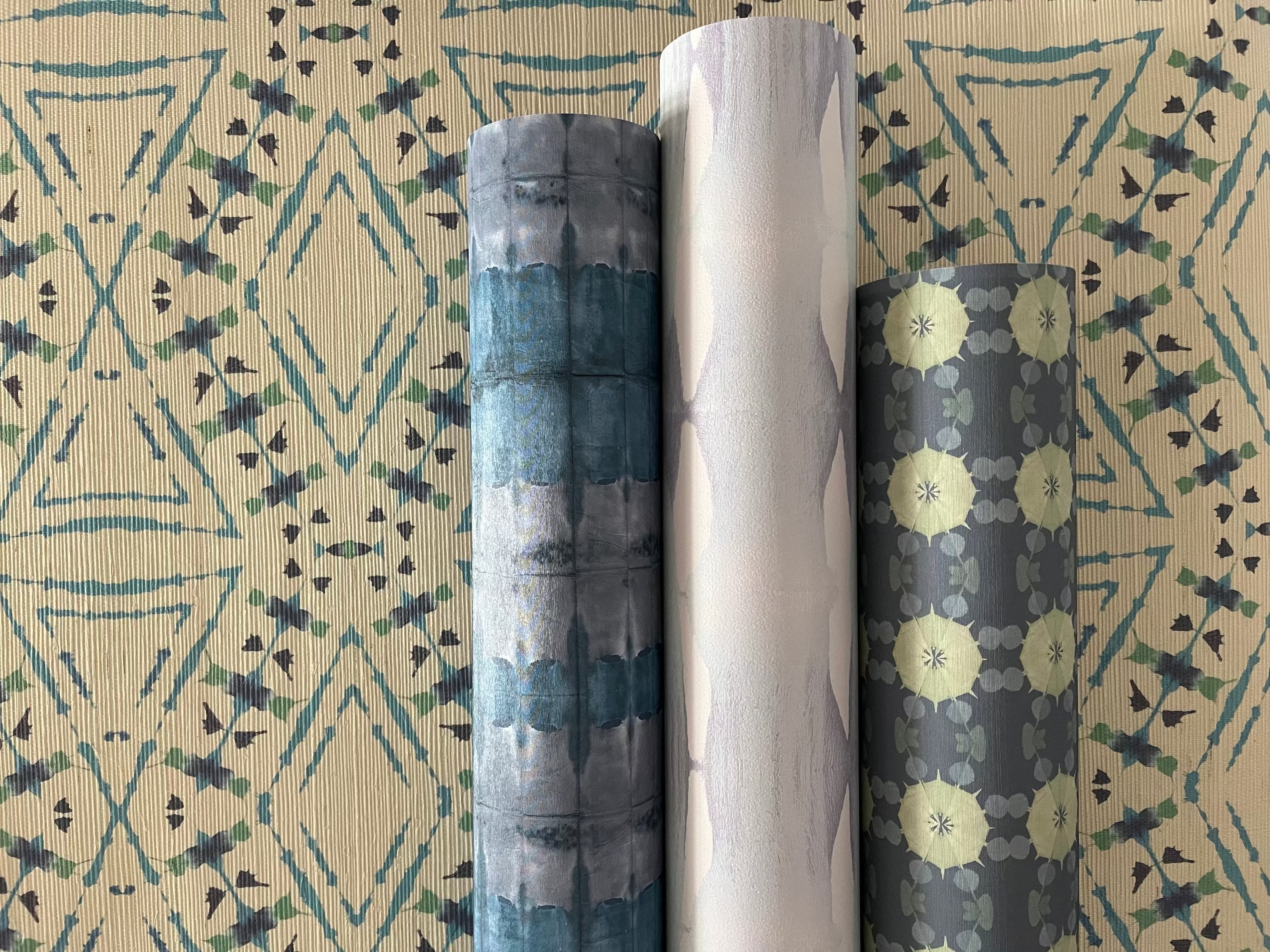 Three rolls of patterned wallpaper on a textured hand-dyed surface with geometric designs.